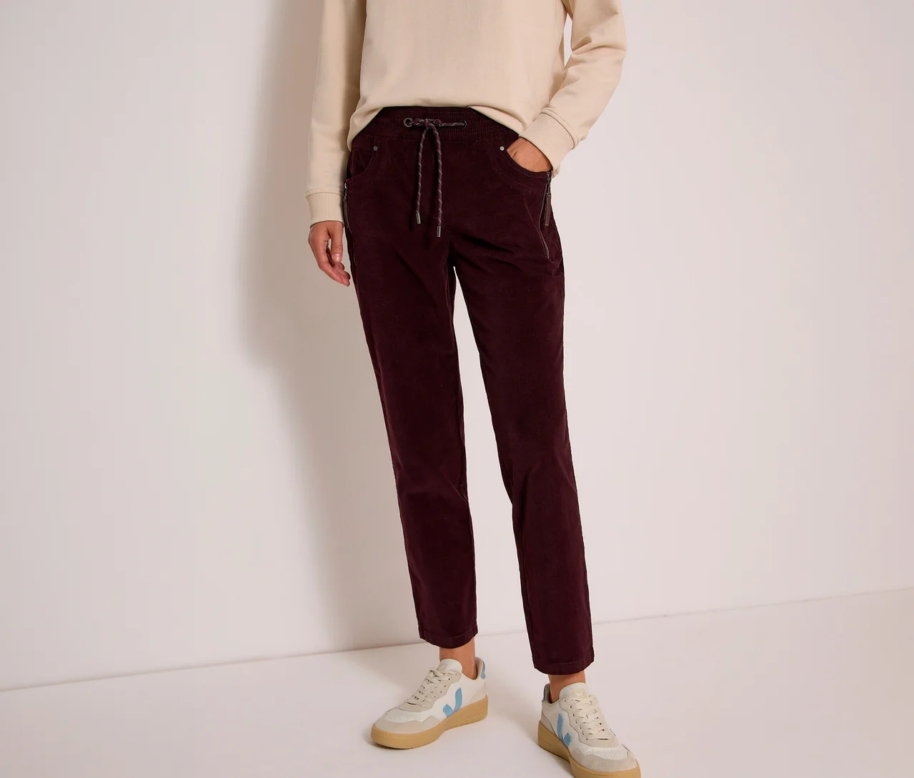 Personne portant un sweatshirt beige et un pantalon en velours côtelé CECIL bordeaux avec cordon de serrage. Des baskets claires sont visibles.