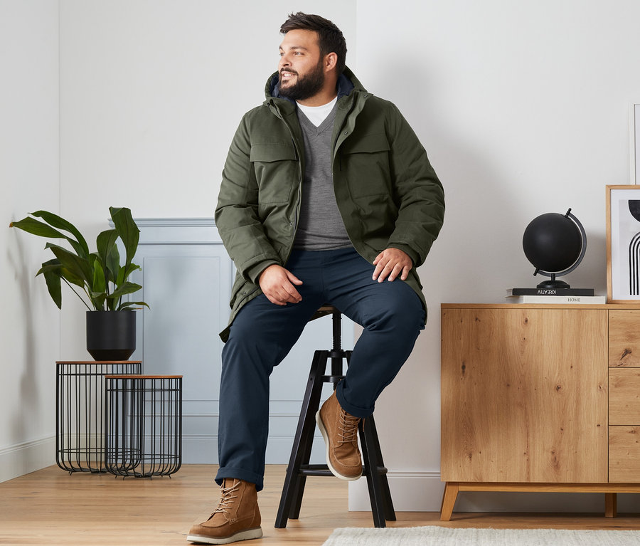 Un homme est assis sur un tabouret et porte une parka vert olive, un pull en laine mérinos gris à encolure en V, un t-shirt blanc et des bottines en cuir velours.