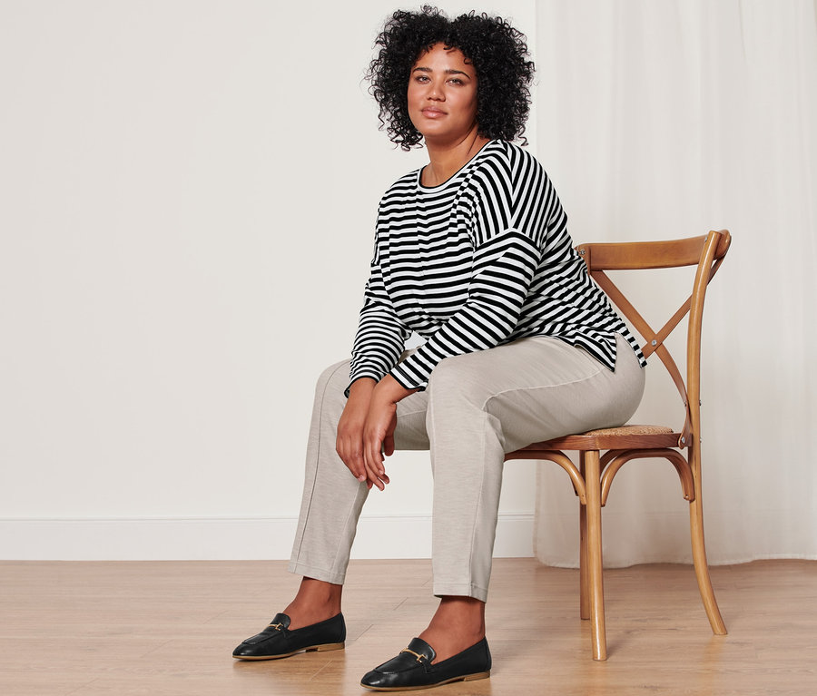 Une femme assise sur une chaise porte un t-shirt à manches longues rayé, un pantalon de jogging beige chiné avec des pinces et des mocassins en cuir.