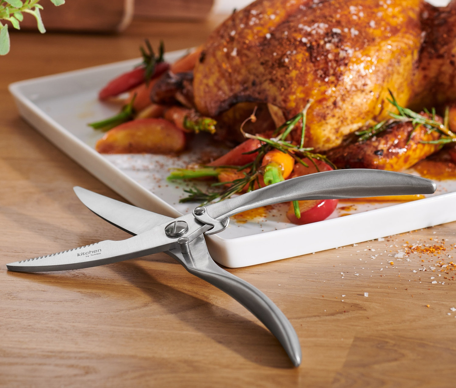 Sur une table en bois reposent un poulet rôti sur une assiette blanche avec des carottes et des herbes, ainsi qu'une cisaille à volaille.