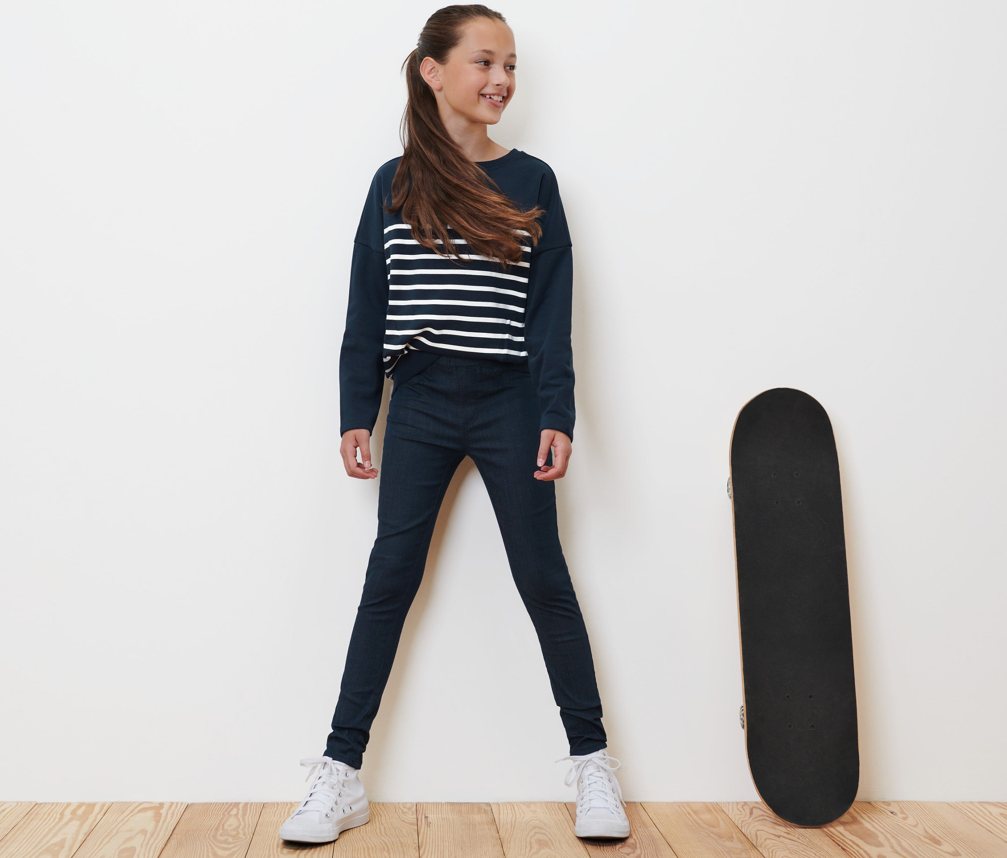 Une fille se tient à côté d'un skateboard et porte un tregging pour enfant – Fit « Jule », bleu.