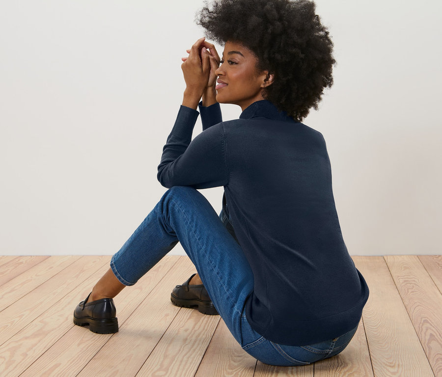 Femme assise sur un plancher en bois portant un pull à col roulé en maille fine bleu foncé.