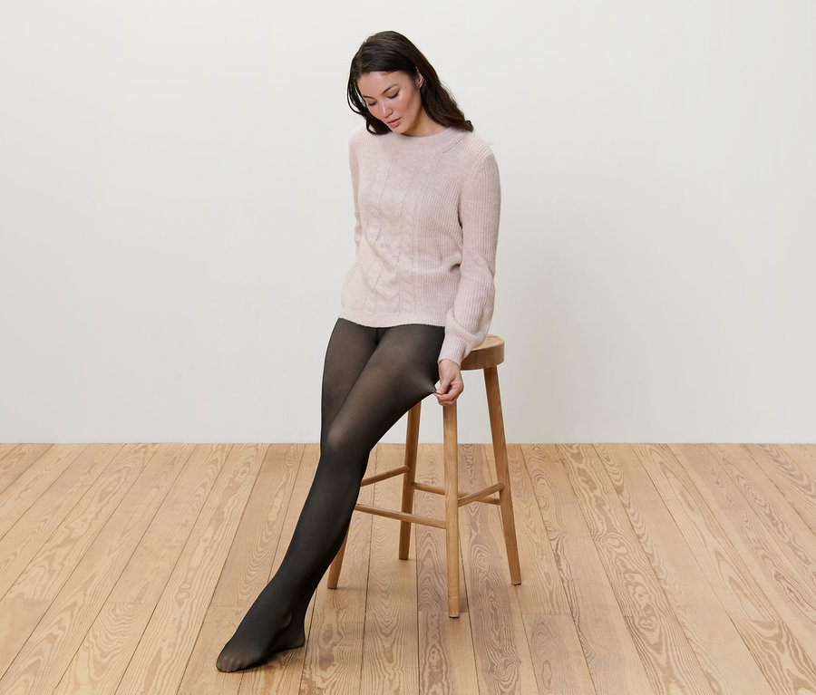 Une femme est assise sur un tabouret et enfile des collants fins doublés en aspect transparent.