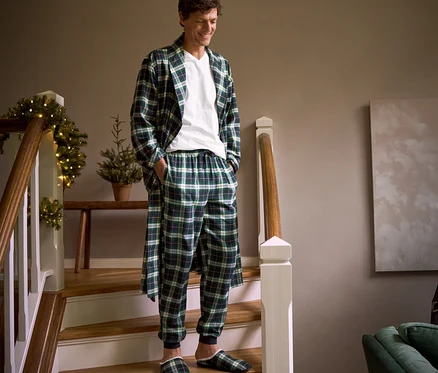 Un homme descend un escalier et porte une robe de chambre en flanelle à carreaux, un pantalon relax en flanelle, des chaussons et 2 t-shirts à encolure en V.