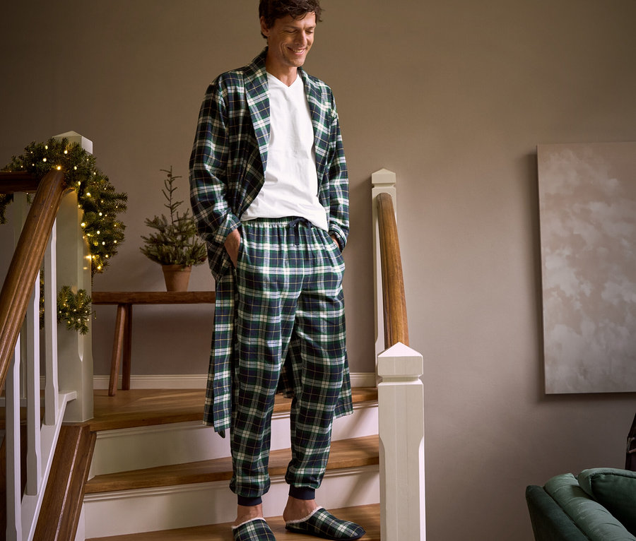 Un homme descend un escalier et porte une robe de chambre en flanelle à carreaux, un pantalon relax en flanelle, des chaussons et 2 t-shirts à encolure en V.