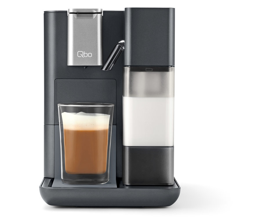 Une machine à café grise Qbo MILK MASTER avec un récipient à lait et un verre rempli de café.