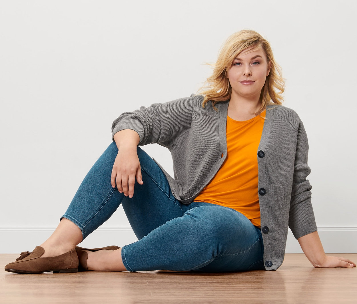 Une femme blonde est assise sur un plancher en bois et porte un tregging «
Fit Nelly
», un chemisier orange, un cardigan en grosse maille et des mocassins en cuir velours.