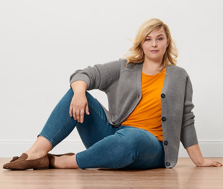 Une femme blonde est assise sur un plancher en bois et porte un tregging «
Fit Nelly
», un chemisier orange, un cardigan en grosse maille et des mocassins en cuir velours.