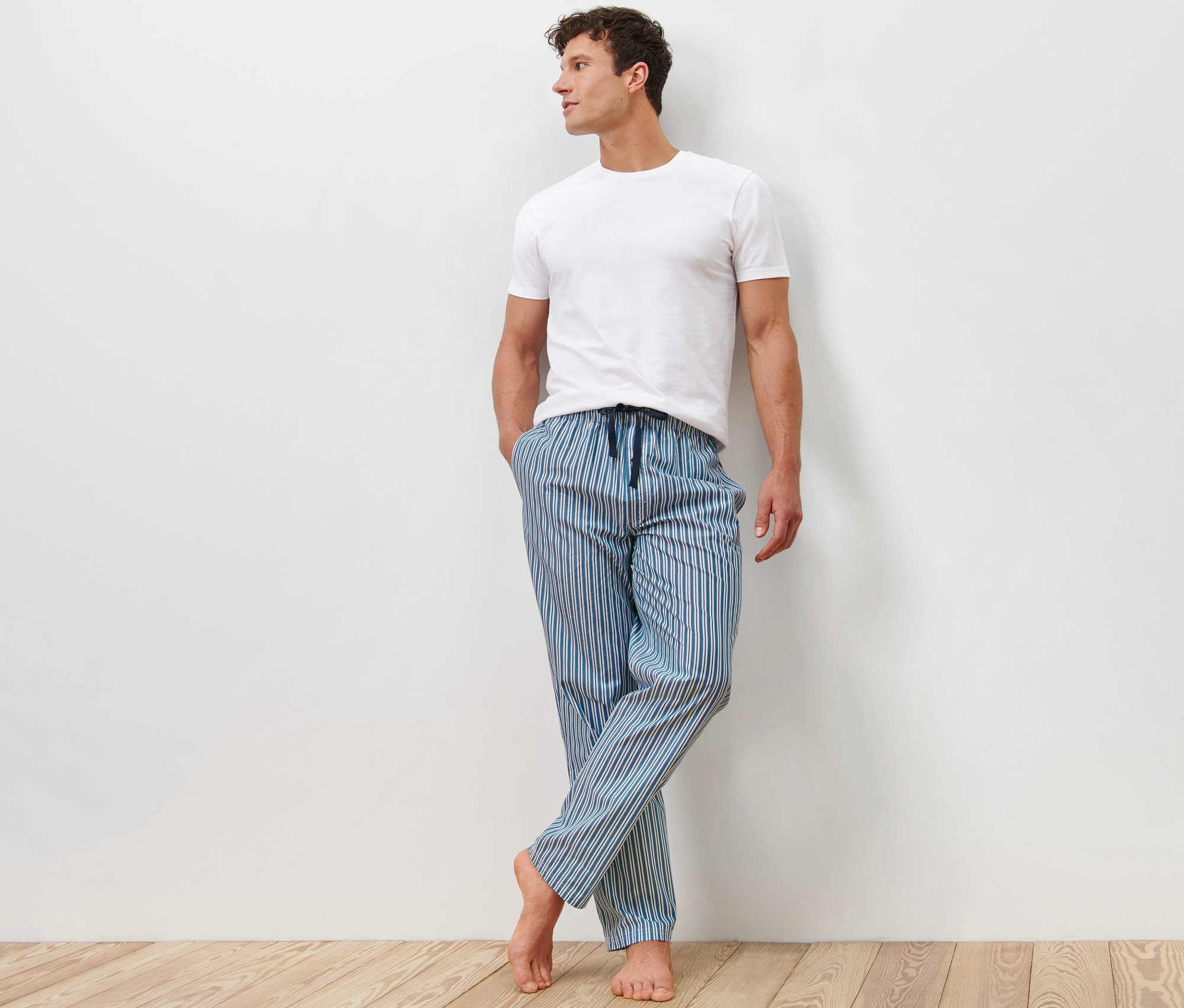 Un homme appuyé contre un mur porte un t-shirt blanc et 2 pantalons de pyjama rayés bleus.