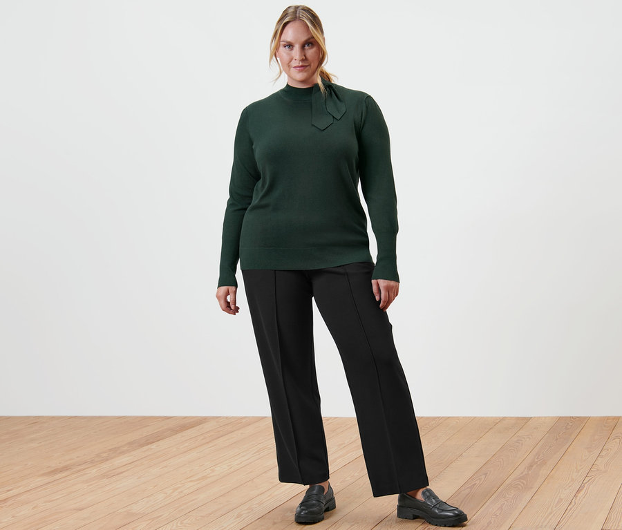 Femme posant en pull en maille fine avec nœud, pantalon en jersey noir avec nervure et loafers en cuir.