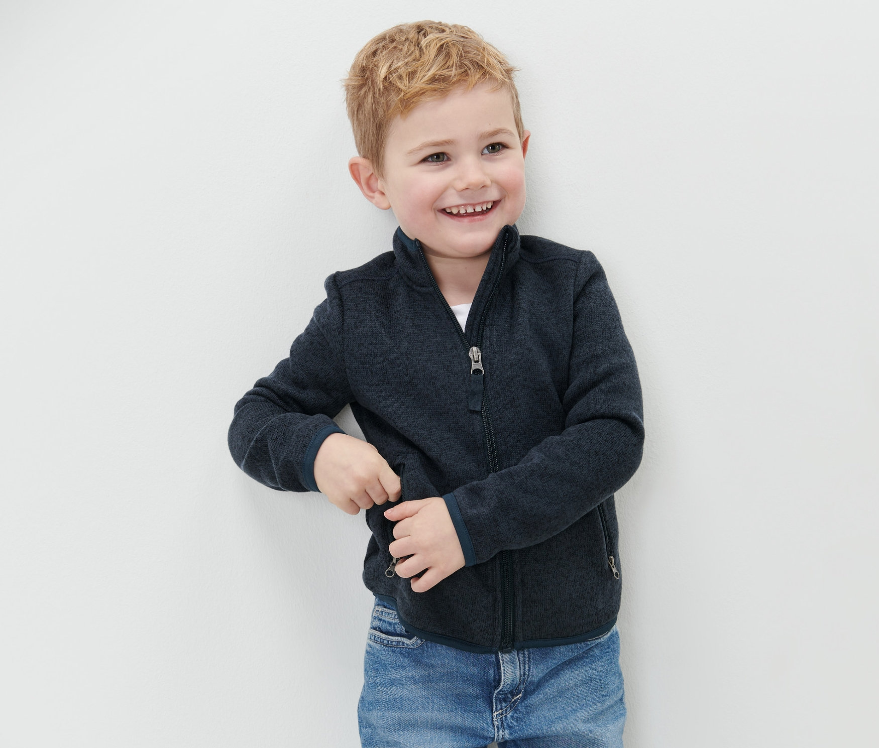 Un garçon souriant se tient près d'un mur et porte une veste en maille polaire pour enfant et un jean bleu.