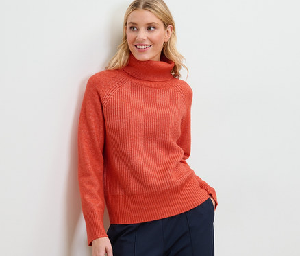 Femme blonde posant en pull à col roulé en grosse maille orange et pantalon bleu marine.
