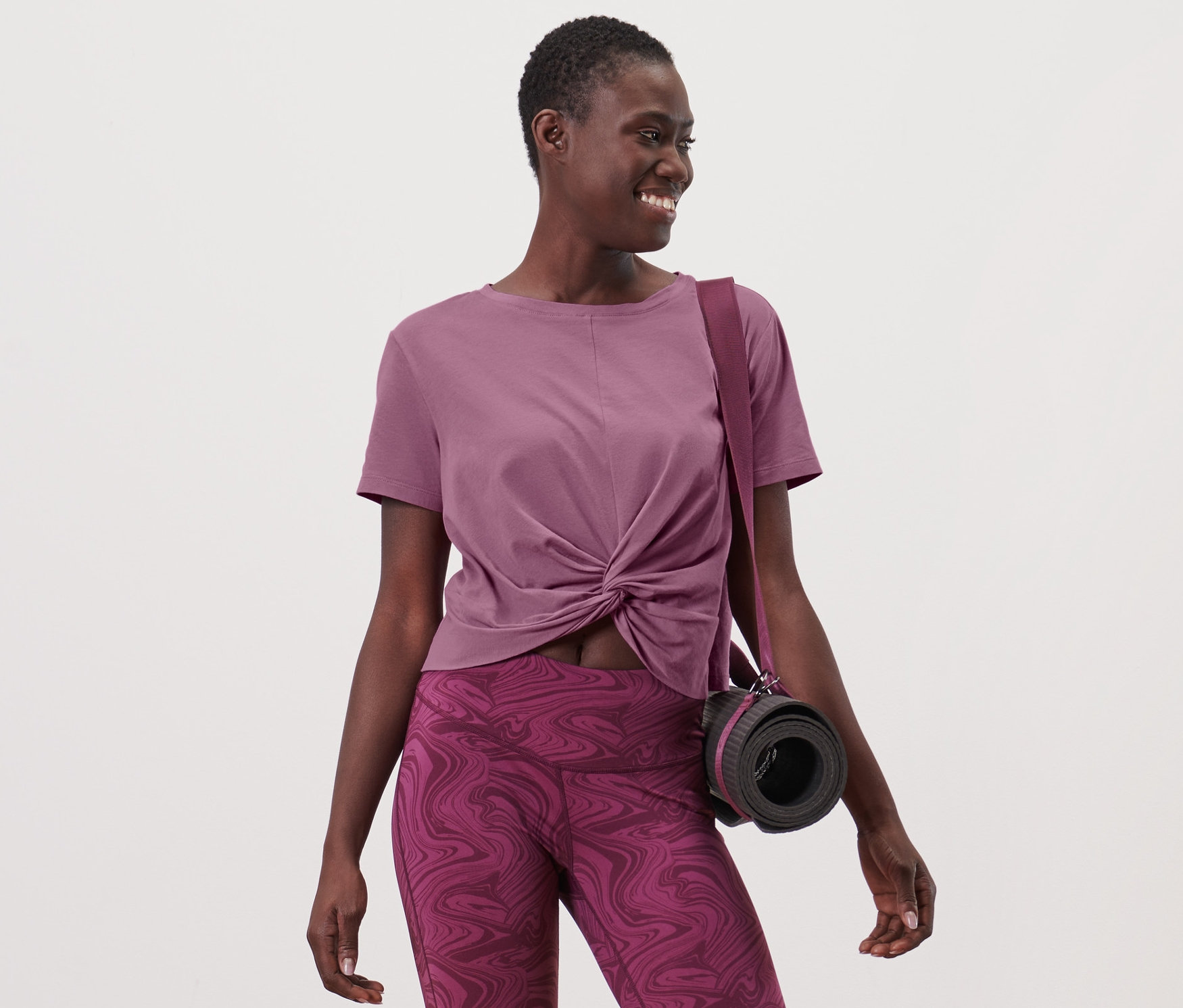 Une femme portant un T-shirt de sport et un legging, avec un sac et un tapis de yoga sur l'épaule.