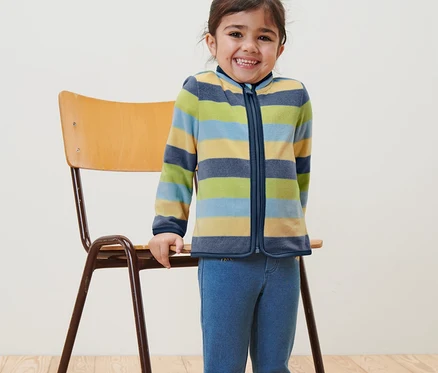 Une fillette souriante se tient à côté d'une chaise et porte une jaquette en polaire pour enfant à rayures.