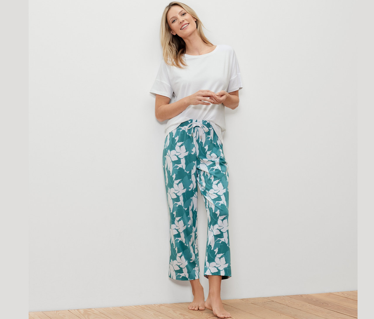 Femme blonde appuyée contre un mur blanc portant un t-shirt blanc et un pantalon de pyjama vert à motifs.