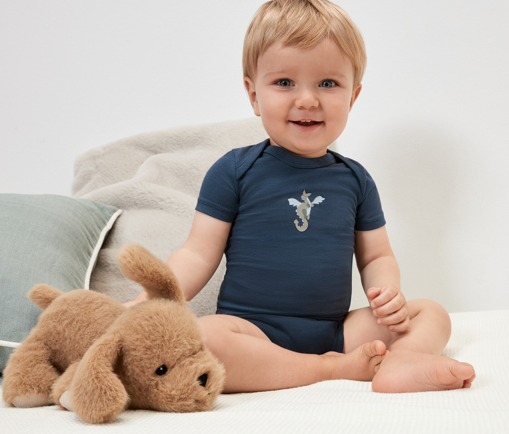 Un bébé souriant est assis avec un chien en peluche et porte un body bleu foncé avec un chevalier.