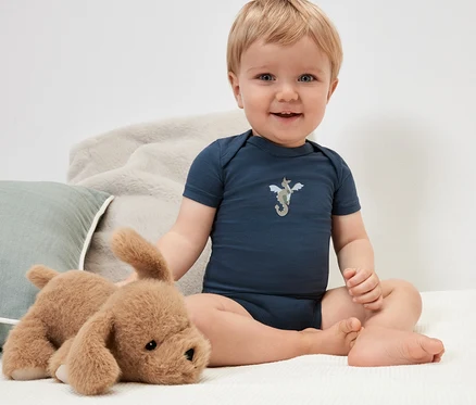 Un bébé souriant est assis avec un chien en peluche et porte un body bleu foncé avec un chevalier.