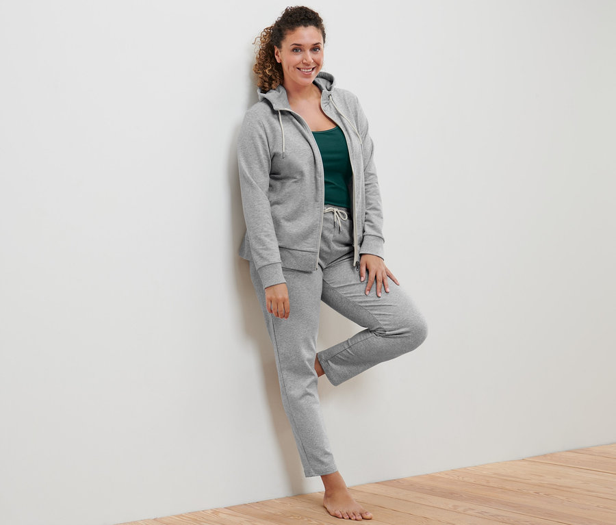 Une femme appuyée contre un mur porte une veste molletonnée grise, 2 tops vert et blanc cassé et un pantalon de jogging gris.