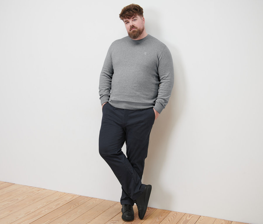 Un homme appuyé contre un mur porte un pull gris à encolure ronde et un pantalon chino bleu marine.