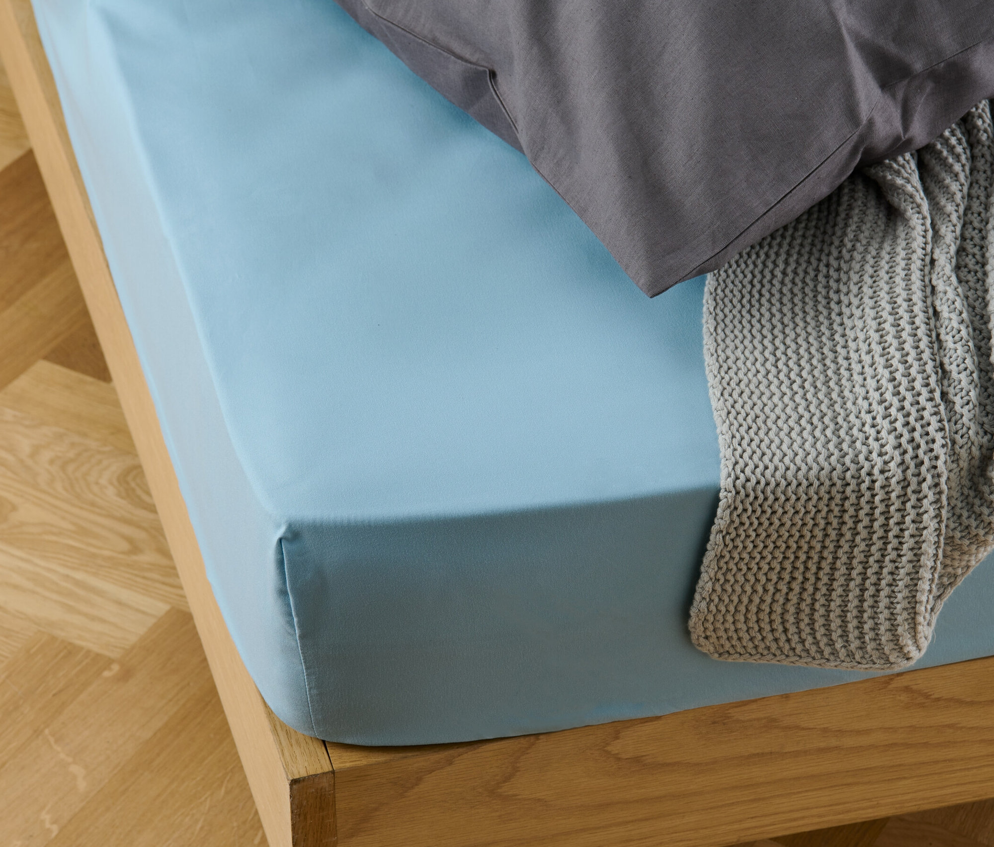 Un drap-housse en microfibres bleu clair sur un lit avec un oreiller gris et une couverture tricotée grise.