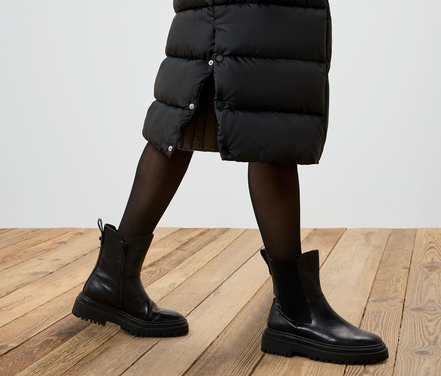 Vue partielle d'une personne portant une jupe noire en duvet, des collants noirs et des bottes en cuir noires sur un plancher en bois.