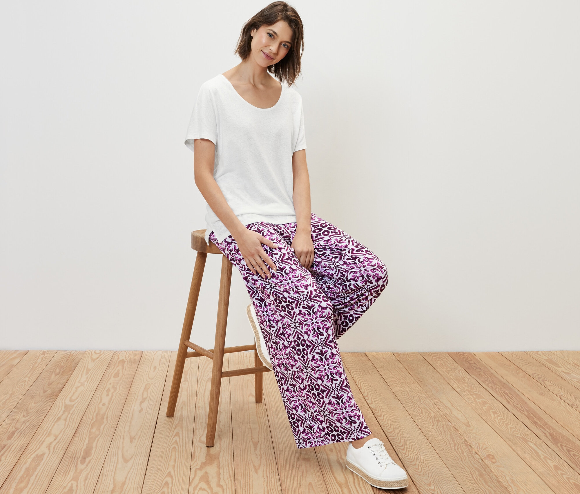 Une femme est assise sur un tabouret et porte un t-shirt blanc en lin mélangé et un pantalon à motifs.