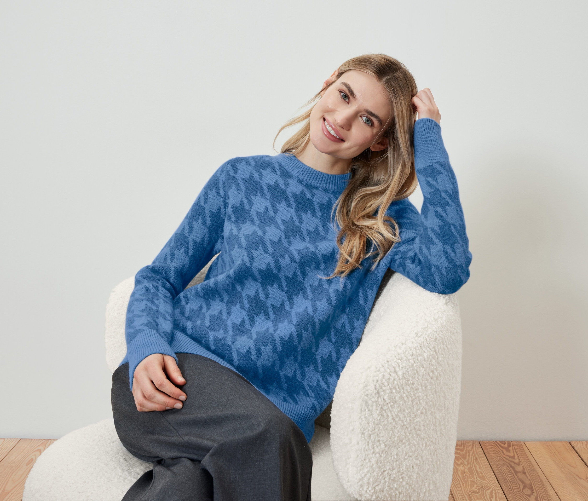 Une femme blonde est assise dans un fauteuil et porte un pull bleu à motifs.