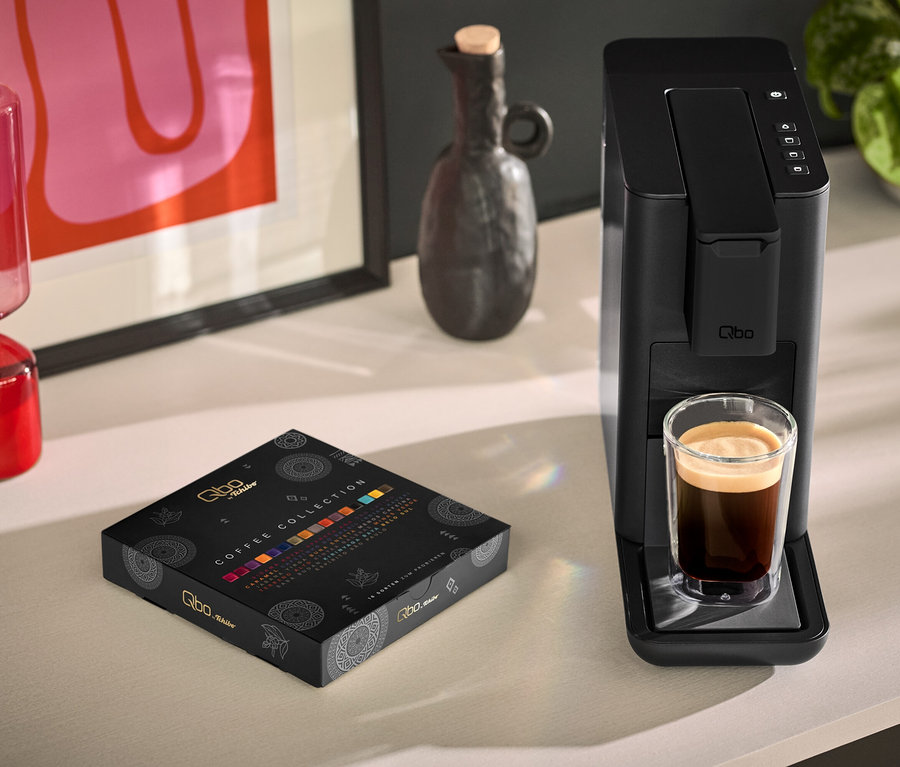Une tasse de café est posée à côté d'une Qbo ICONIC Dark Chrome noire et d'un paquet de Qbo Collection - 16 capsules.