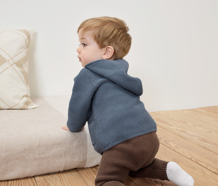 Bébé rampant sur un canapé beige vêtu d'un sweat à capuche bleu et d'un pantalon marron.