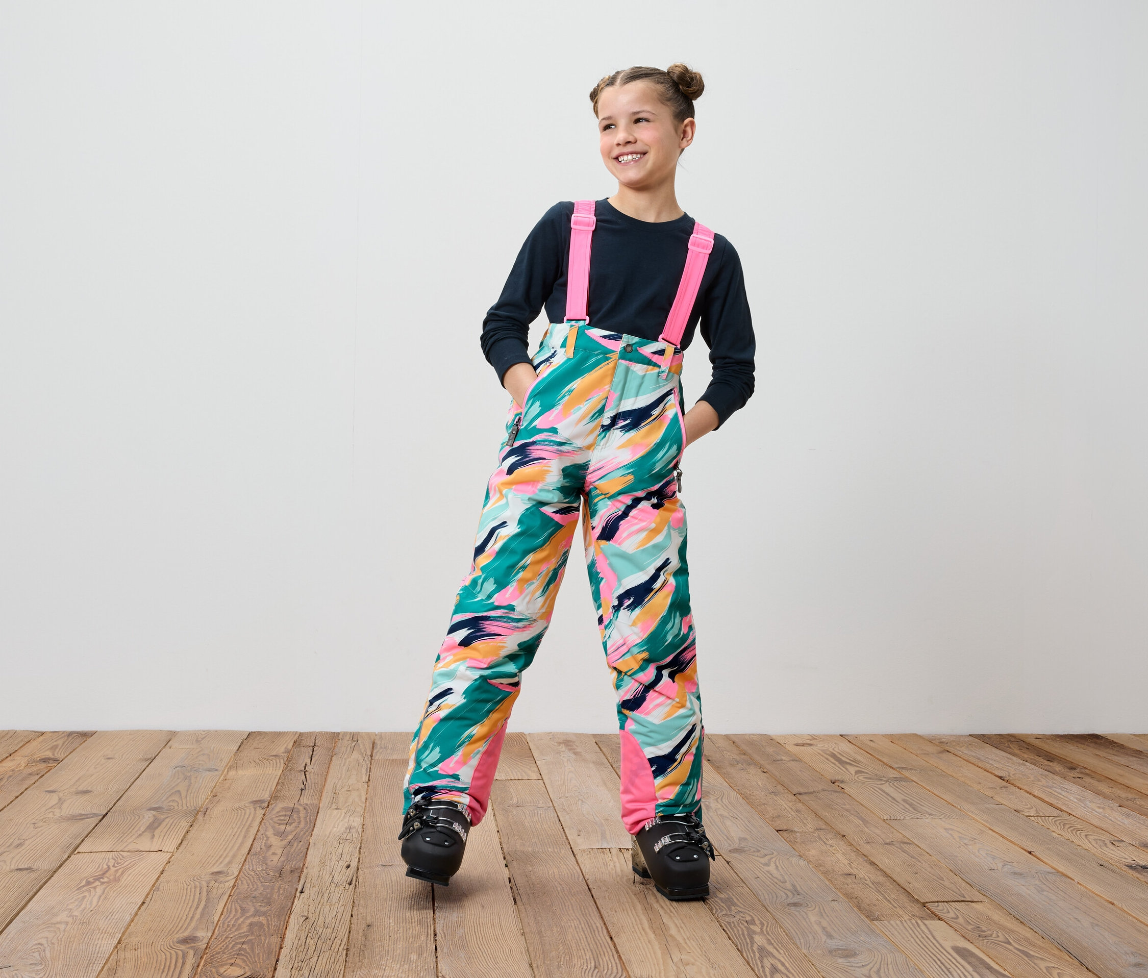 Adolescente souriante portant un pantalon de ski coloré à bretelles et des chaussures de ski noires, debout sur un plancher en bois.