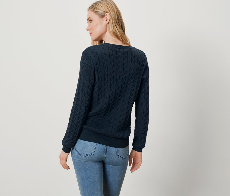 Une femme blonde, vêtue d'un pull bleu et d'un jean, se tient dos à l'appareil.