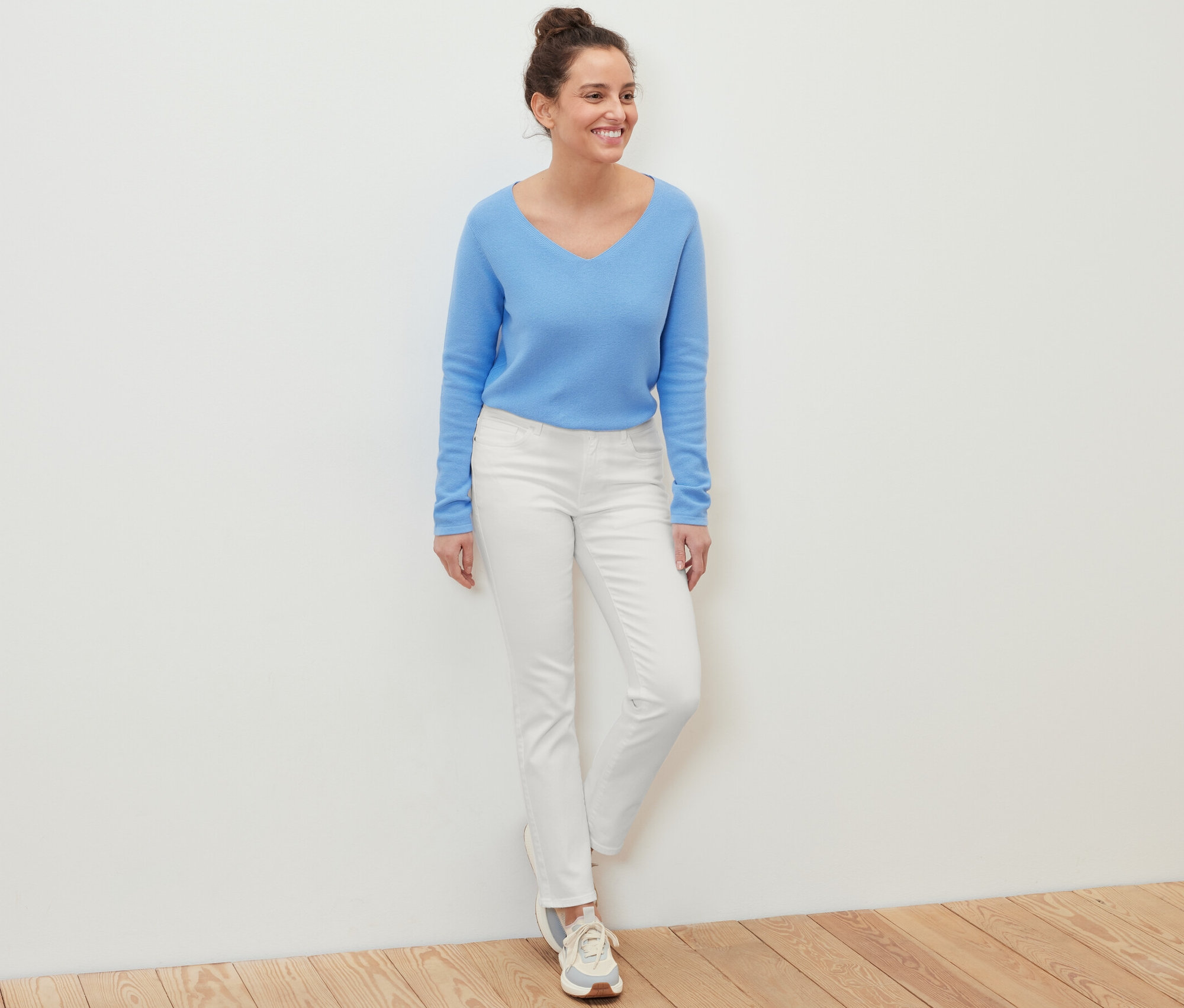 Une femme debout souriant, portant un pull bleu et un jean « Kira » blanc.