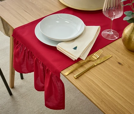 Table dressée avec une nappe rouge, de la vaisselle blanche, une serviette beige avec un motif d'arbre de Noël, des couverts dorés, un verre à vin et un vase doré avec de l'eucalyptus.