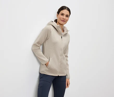 Une femme se tient debout, la main dans la poche d'un sweat à capuche beige zippé et d'un pantalon bleu.