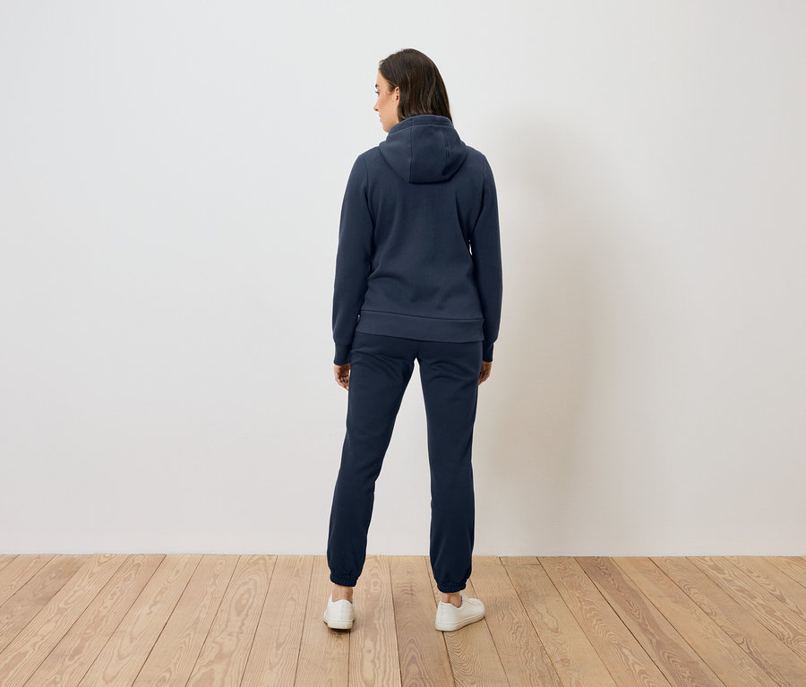 Une femme vue de dos portant un sweat à capuche bleu marine et un pantalon de survêtement, se tenant sur un plancher en bois devant un mur blanc.