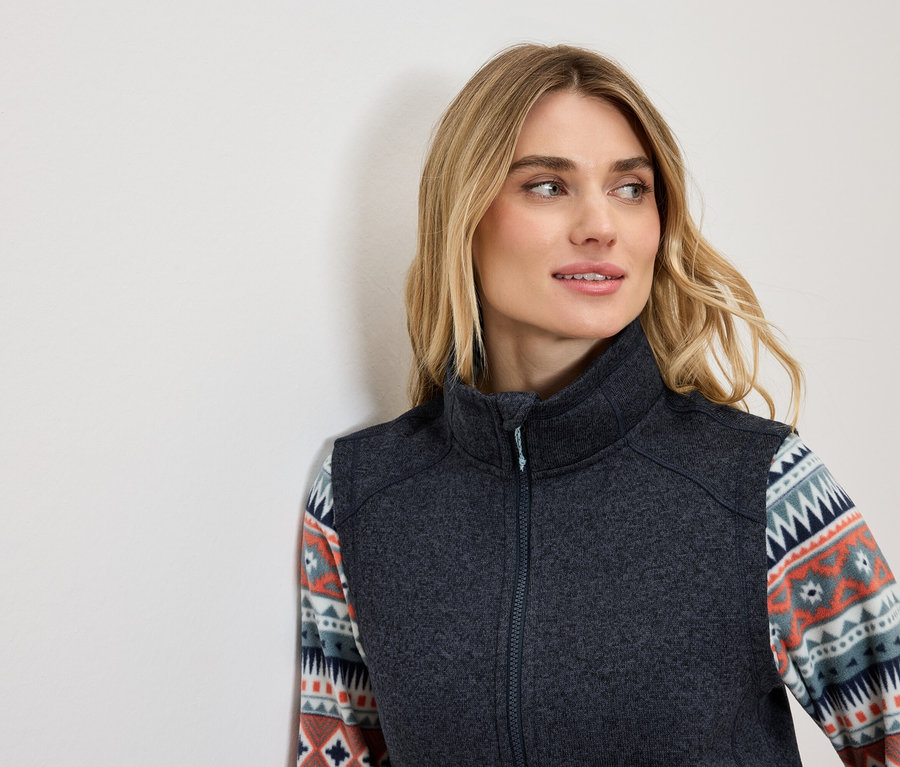 Blonde portant un gilet gris foncé avec un motif décoratif sur les manches, regardant vers la gauche.