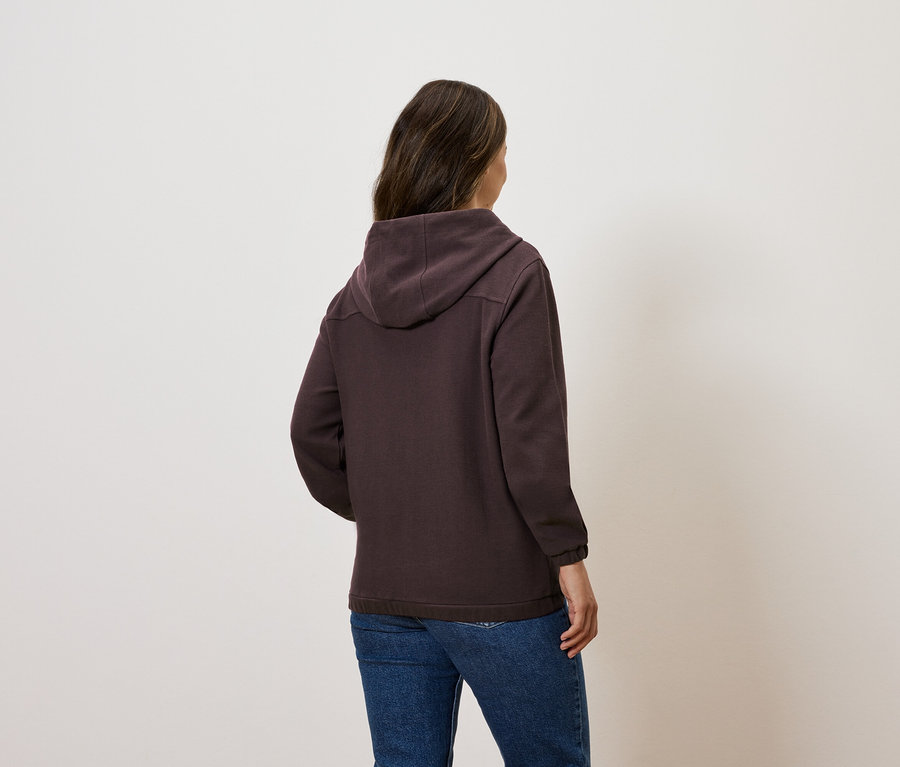 Une femme se tient dos à la caméra et porte un sweat à capuche marron et un jean.