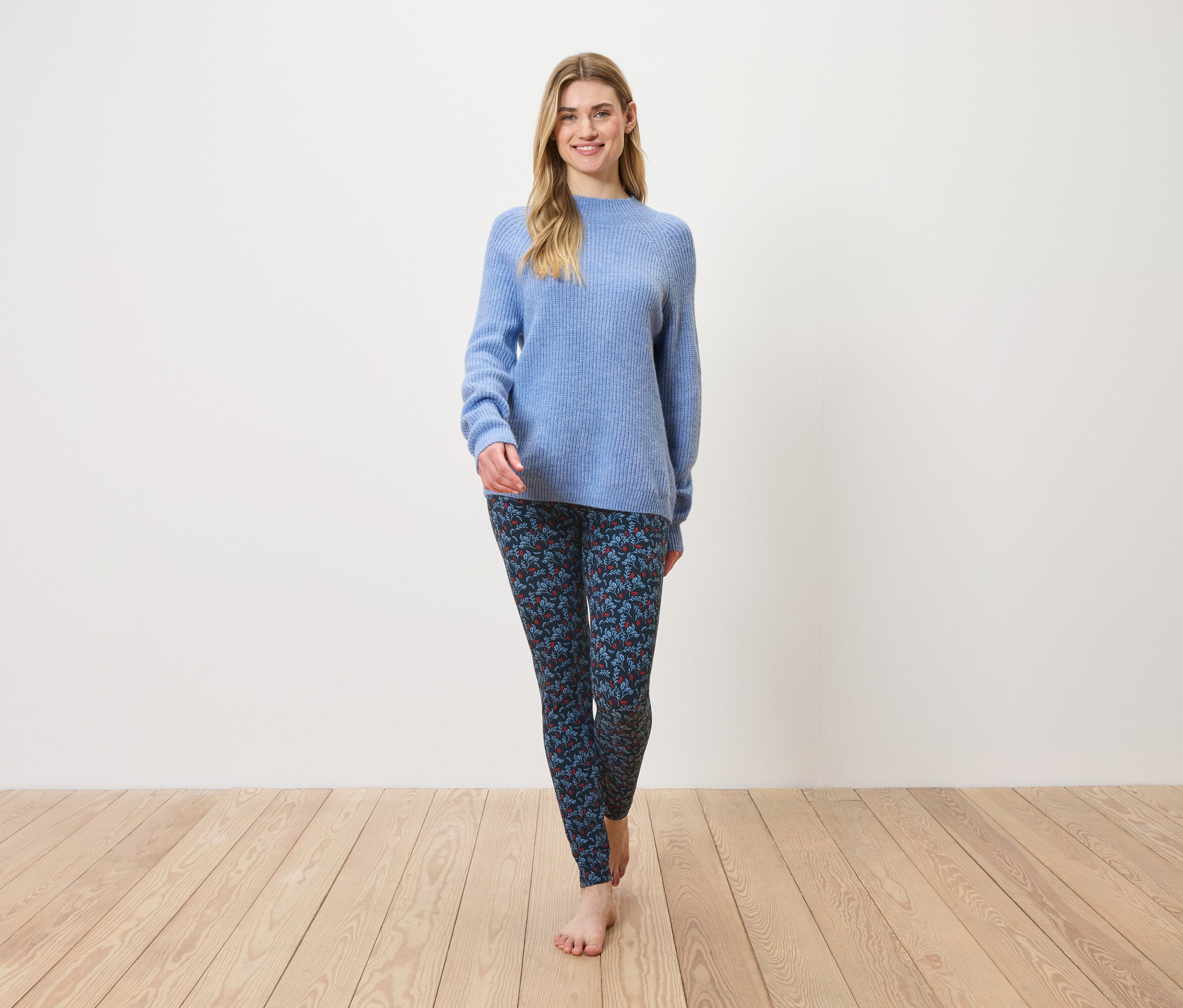 Une femme marche pieds nus sur un plancher en bois, portant un pull bleu et un legging à motifs.