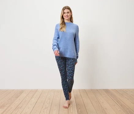 Une femme marche pieds nus sur un plancher en bois, portant un pull bleu et un legging à motifs.