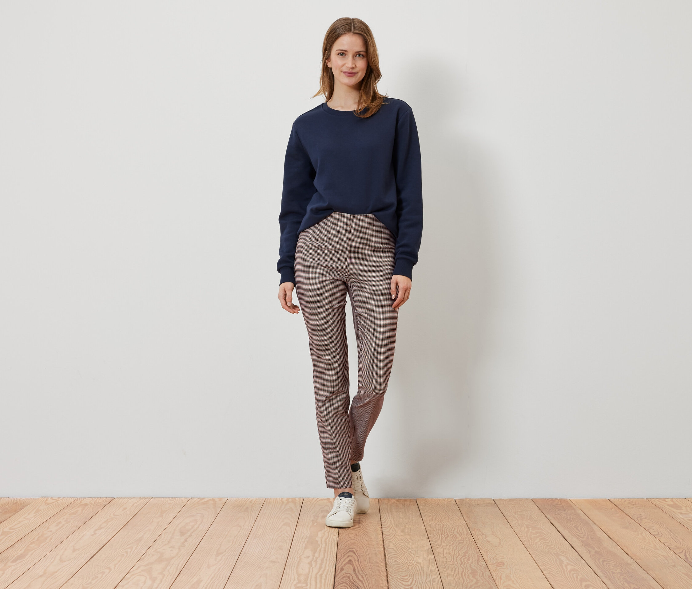 Une femme se tient sur un plancher en bois, portant un sweat-shirt bleu marine et un pantalon à carreaux marron.