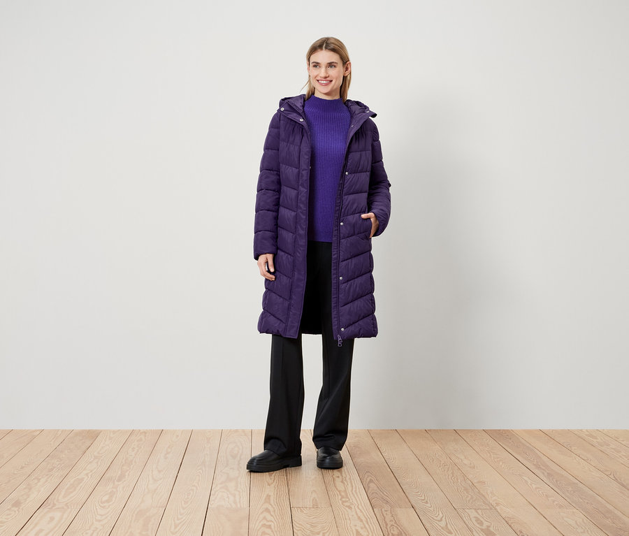 Une femme se tient sur un plancher en bois et porte un manteau violet, un pull violet et un pantalon noir.