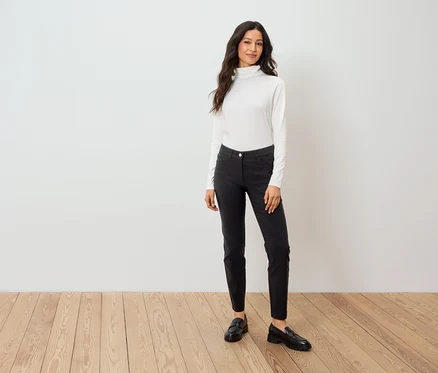 Une femme se tient sur un plancher en bois, portant un pull à col roulé blanc, un pantalon noir et des mocassins noirs.
