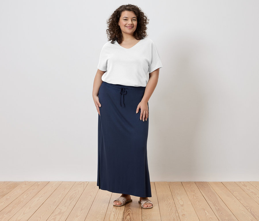 Une femme debout portant un t-shirt blanc, une longue jupe bleu marine et des sandales.