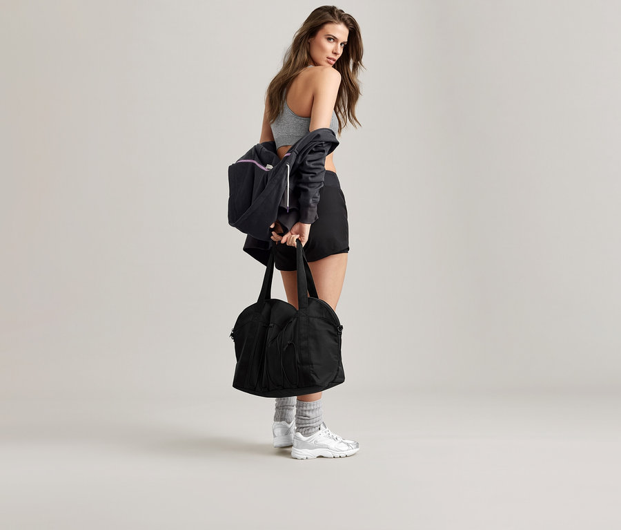 Une femme posant sur un fond neutre, portant une veste de survêtement anthracite, un soutien-gorge de sport, un short, des chaussettes et des baskets, tenant un sac de sport noir.