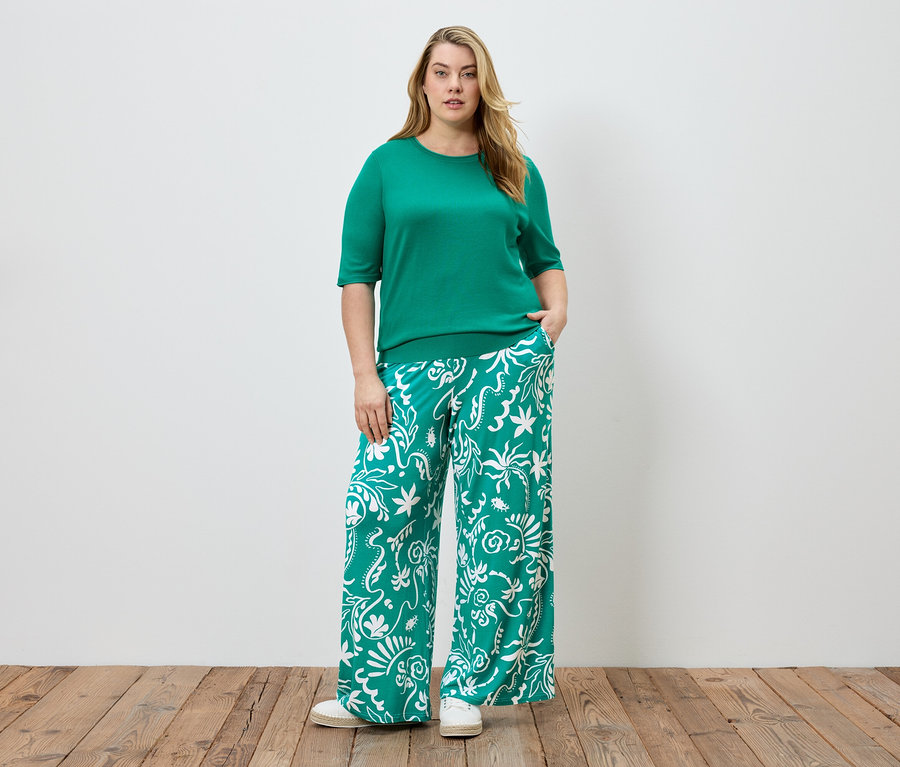 Une femme se tient devant un mur blanc, portant un haut vert et un pantalon vert à motifs blancs.
