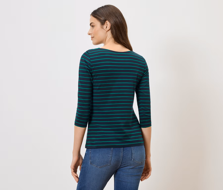 Une femme brune, vue de dos, porte un t-shirt rayé bleu et vert et un jean sur fond blanc.