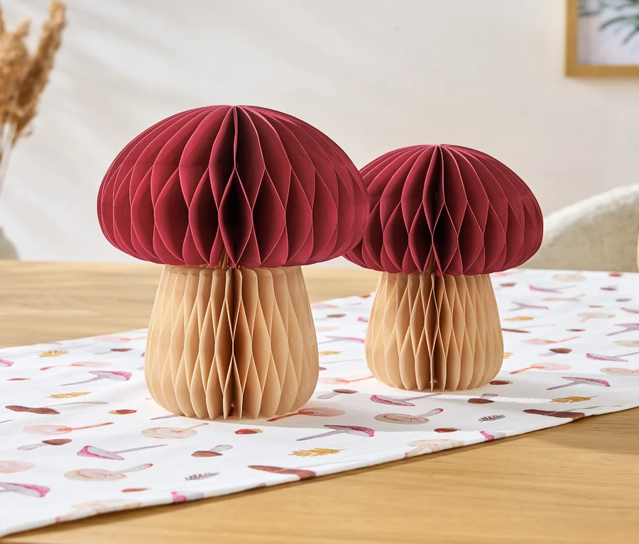 Deux champignons décoratifs avec des têtes rouges et des pieds beiges sont posés sur un chemin de table à motifs de champignons.