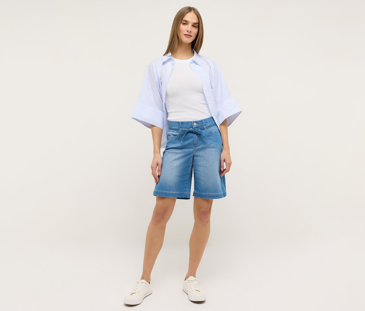 Une femme porte un short Angels Denim « Lia City » bleu clair, un t-shirt blanc, une chemise rayée bleue et des baskets blanches.