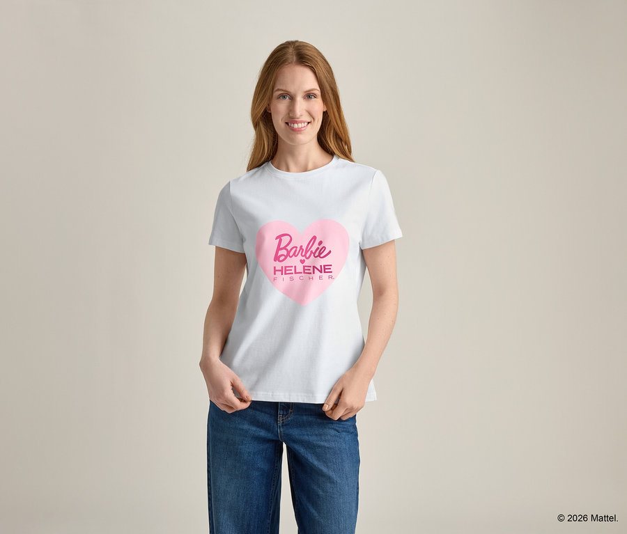 Une femme aux cheveux blonds porte un t-shirt blanc avec un imprimé cœur rose et l'inscription "Barbie HELENE FISCHER", ainsi qu'un jean bleu.