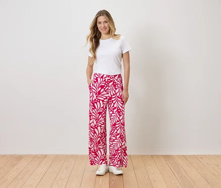 Une femme portant un t-shirt blanc et un pantalon rouge et blanc à imprimé floral se tient devant un mur, une main dans la poche.